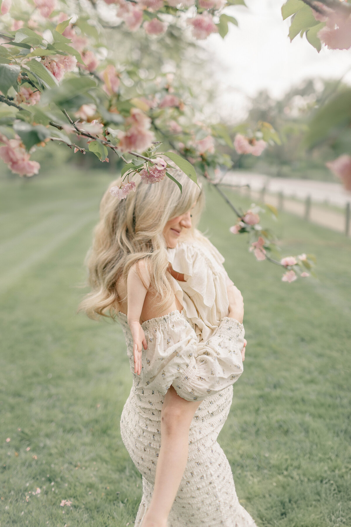Pregnant mother lifting her daughter under a blooming tree, both wearing soft dresses, during an outdoor maternity session in a Kansas City park.