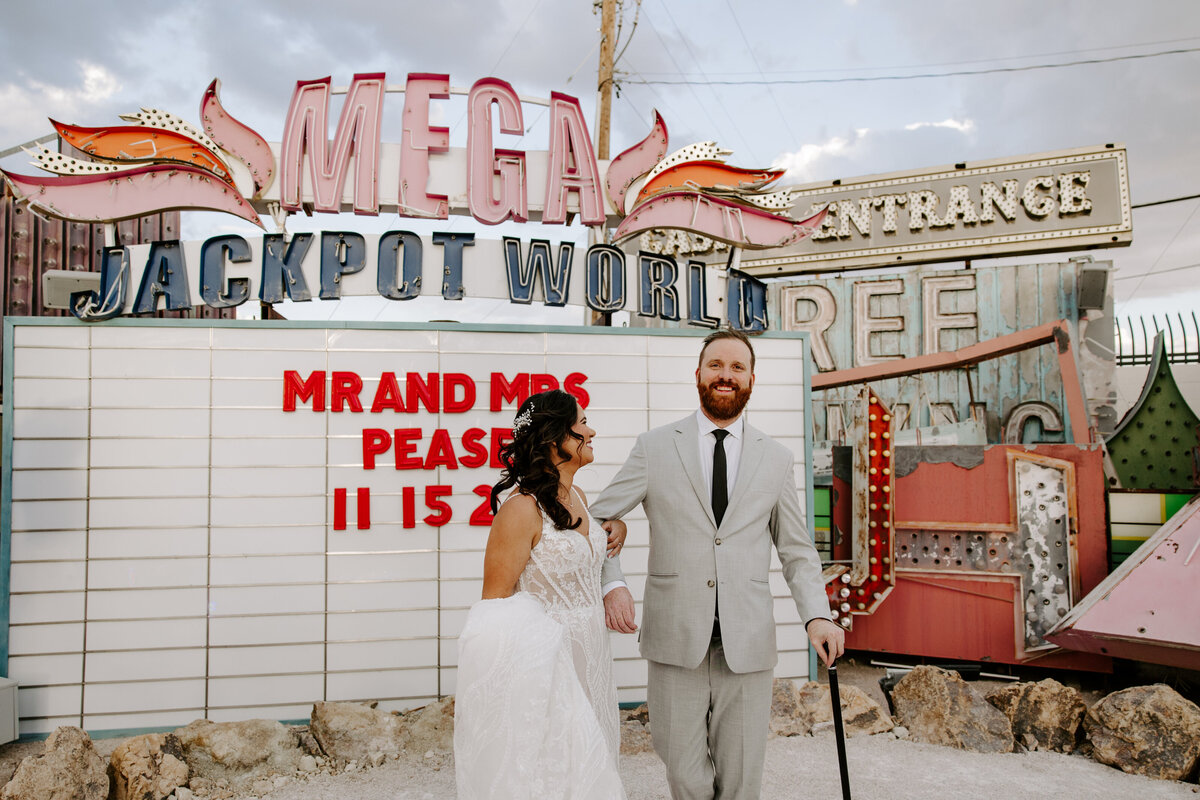 A couple smiles and poses in front of neon signs.
