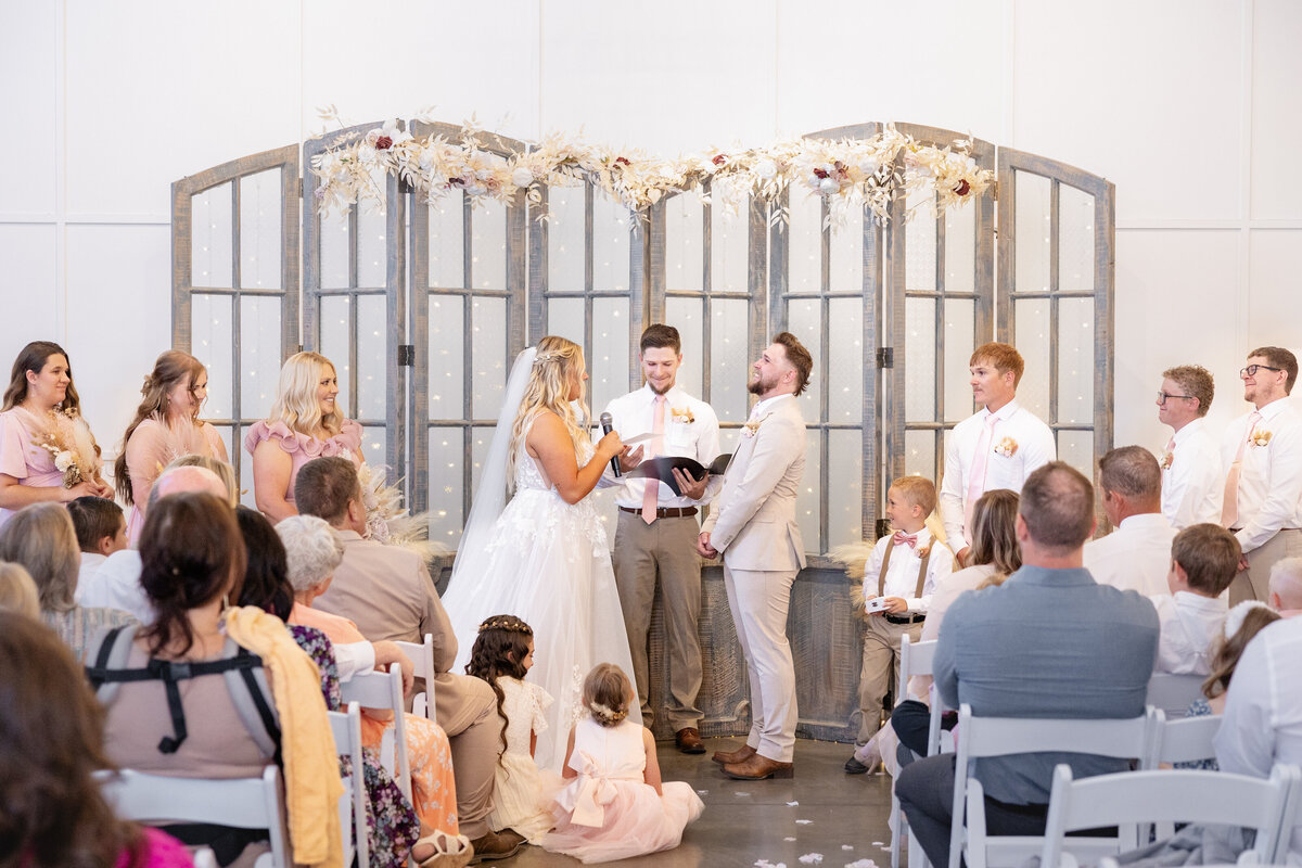 The groom laughs as the bride shares her vows, bringing warmth and lightheartedness to the ceremony. The wedding officiant smiles, and guests look on, celebrating the couple’s love. Captured at Maplewood Events in Spanish Fork, Utah by Cassie Lee Photography.