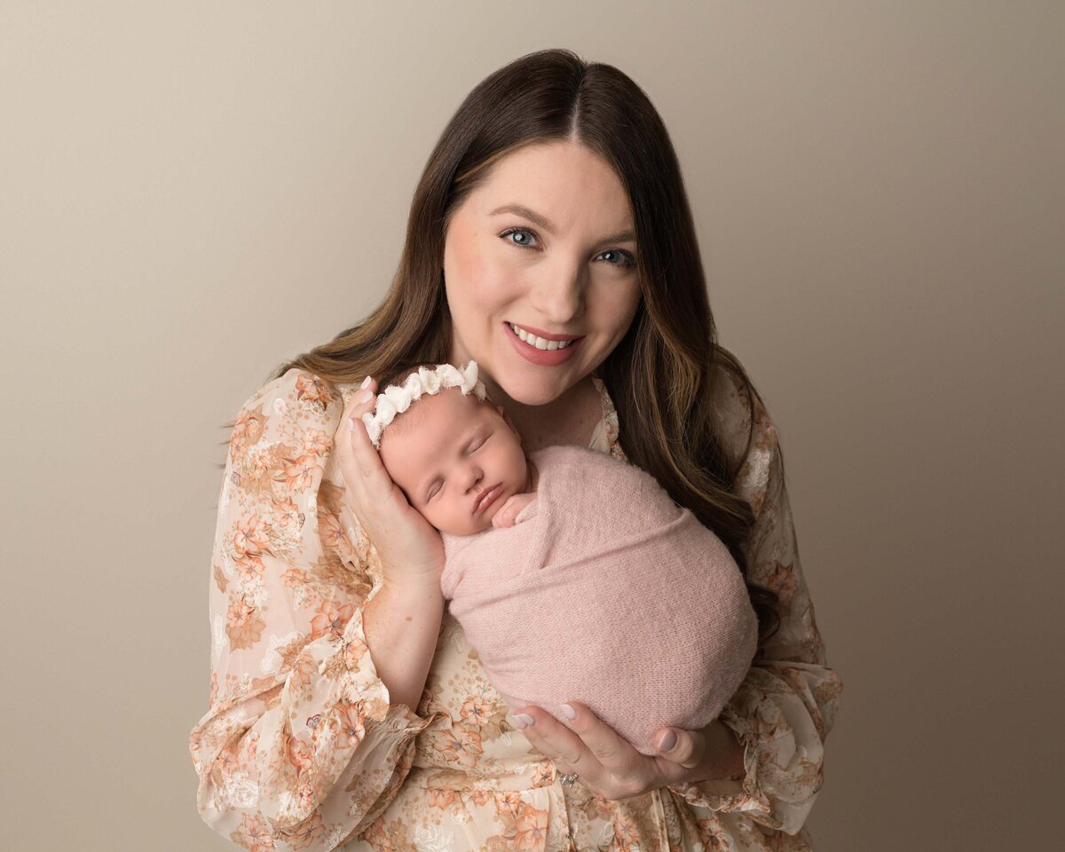 Mom poses for a photo with her baby girl during her Lake Elsinore newborn photoshoot. Mom is holding baby, swaddled in pink, so she is facing the camera. Baby is sleeping and mom is smiling at the camera. Captured by best Lake Elsinore newborn photographer Bonny Lynn Photography.