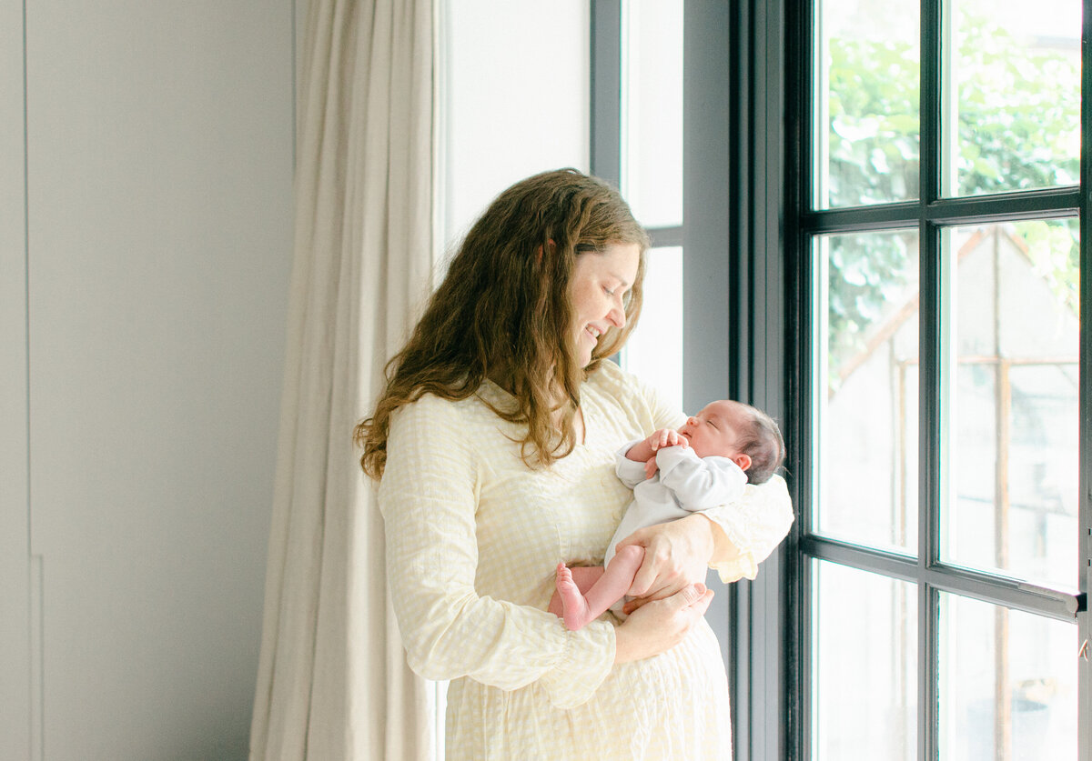 Mother in a yellow checkered dress cradles her newborn dressed in white by the window of their home in Amsterdam.