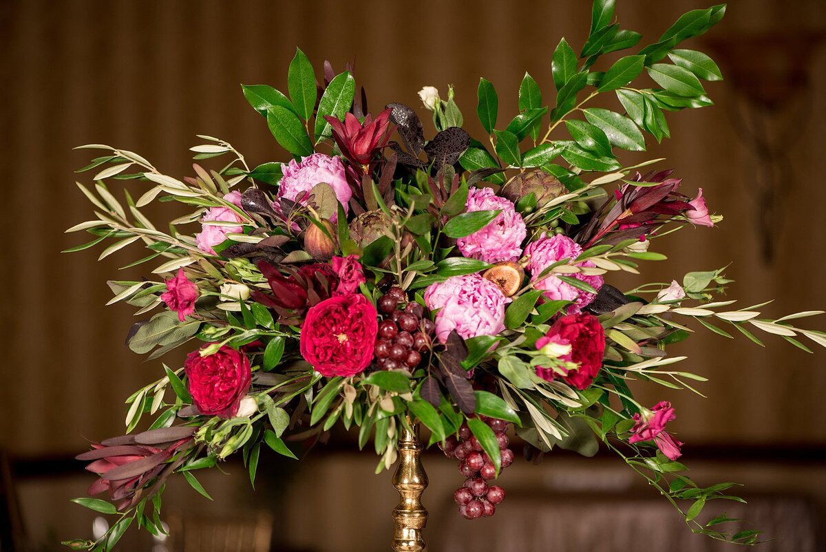 A large floral centerpiece on a thin gold stand featuring a lush selection of red peonies, pink peonies, red roses, pink roses, red protea, pink protea, artichokes, dangling clusters of grapes and assorted greenery at Embassy Suites Hotel wedding.