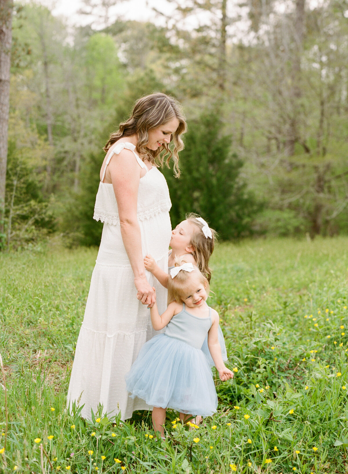 Sisters hold mom's hand and kiss her belly. Image by Raleigh maternity photographer A.J. Dunlap Photography.