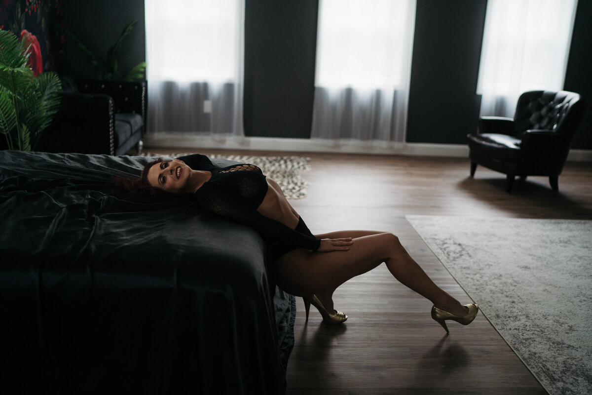 A woman in black lace lingerie leans back over a bed with a hand on her leg in a Boston boudoir photography session