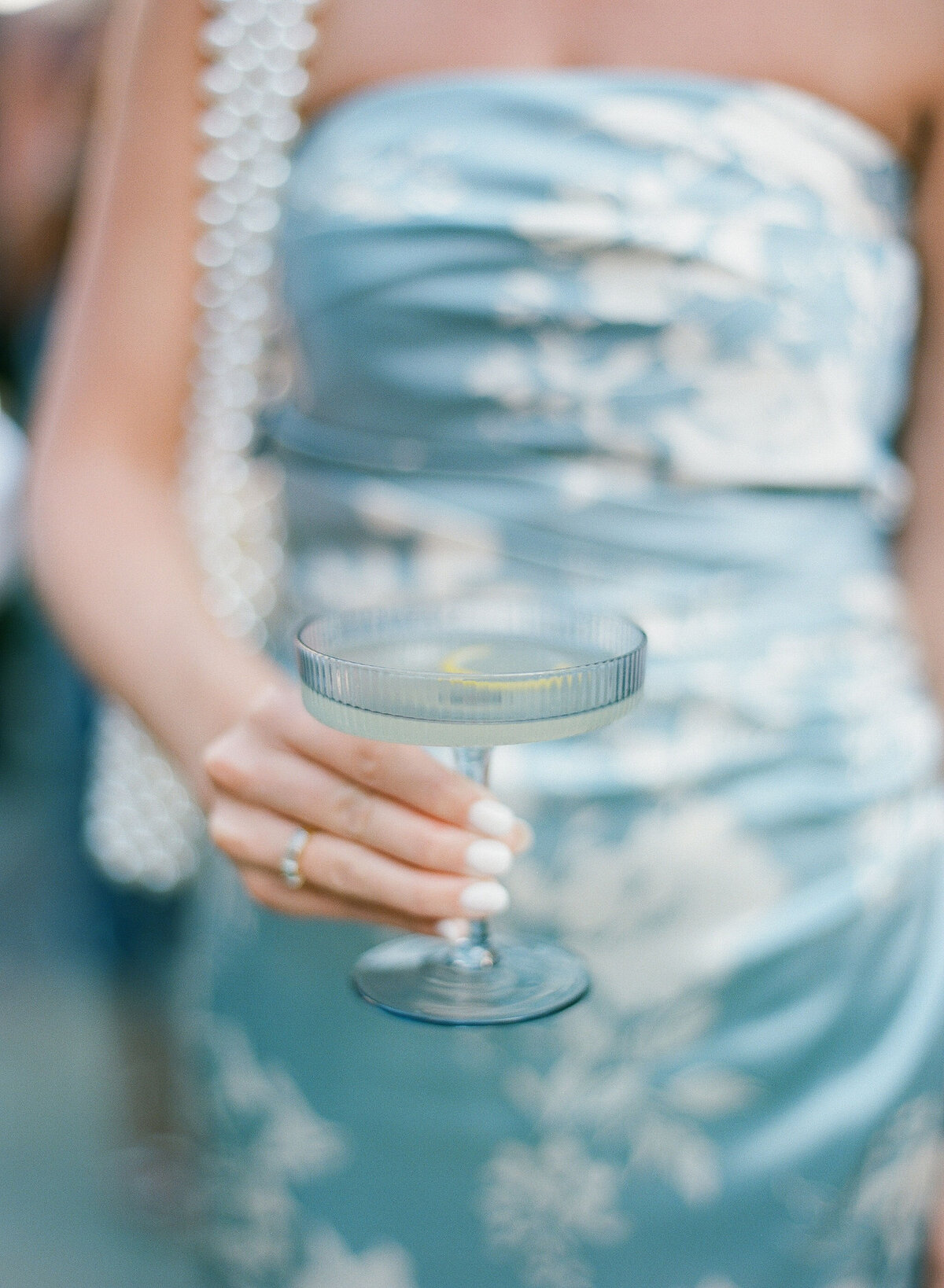 This photo was taken at Washington National Cathedral in Washington D.C. by photographer Twah Dougherty. In the image, a person wearing a strapless blue and white floral dress can be seen holding a delicate glass of champagne. The focus is on the glass and the person's hand, showcasing white painted nails and a ring on the finger. Additionally, a pearl strap is visible over the shoulder, adding a touch of elegance to the overall composition.