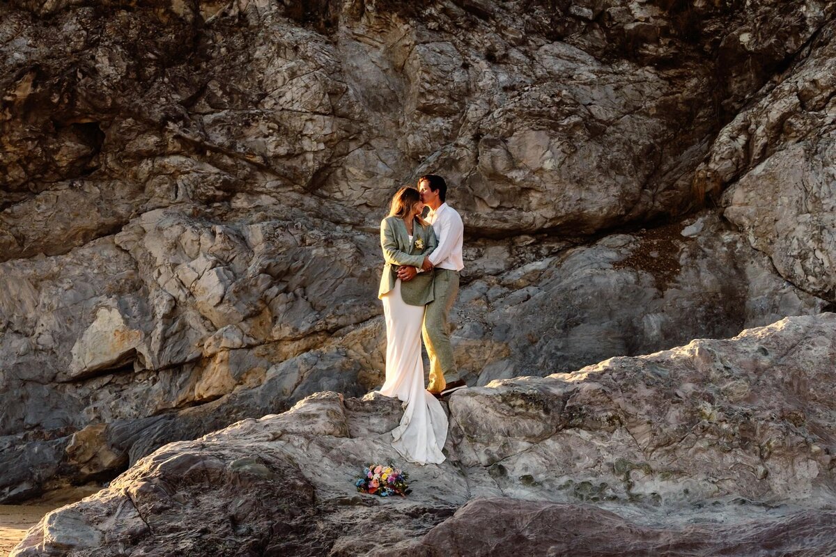 during their Oregon coast elopement, a couple stands on a coastal rock in their wedding attire. The groom kisses the bride on her forehead as she bundles up in his jacket