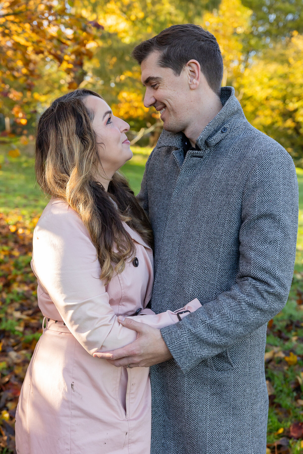 A couple embracing and smiling at each other outdoors with trees showing autumn colors in the background.