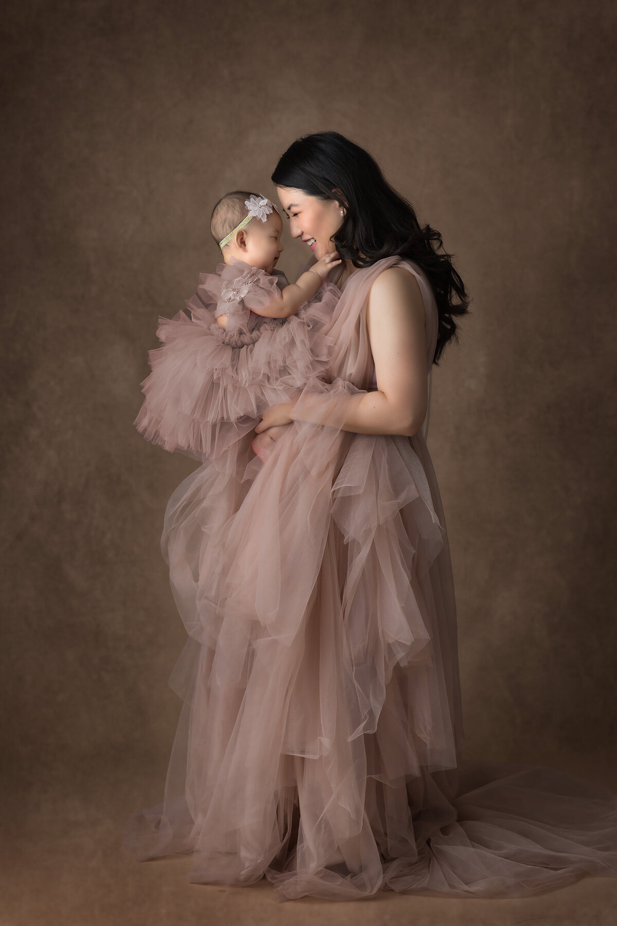 A mother and her baby in soft pink tulle gowns. This family photography in Dallas showcases a loving, joyful moment against a neutral backdrop. The image highlights the bond between mother and child.