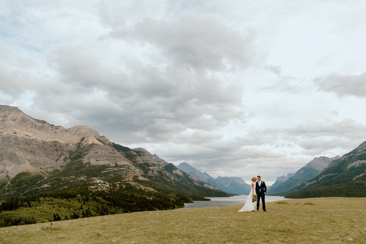 Couple standing on top of a hill in Waterton, AB near the Prince of Whales Hotel, captured by Love and be Loved Photography, authentic and natural wedding photographer and videographer in Lethbridge, Alberta. Featured on the Bronte Bride Vendor Guide.
