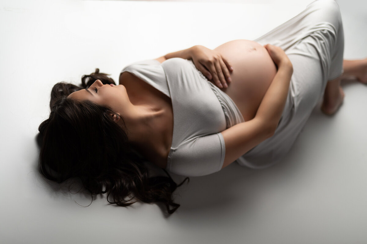 Pregnant woman in a soft gray gown lying on a seamless white studio floor, gently holding her belly with eyes closed during a peaceful, minimalist maternity session in Waukesha near Hartland, Wisconsin.
