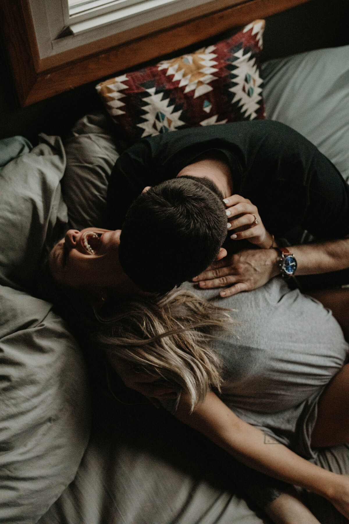 A couple lies in bed, with the man leaning down to kiss the woman, who is laughing heartily.