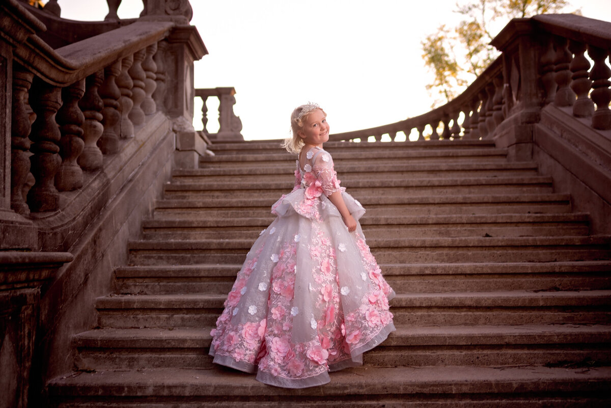 Elegant portrait of a young girl in a flowing pink gown standing on grand stone stairs at sunset. Captured in Leawood, Kansas, this regal scene is ideal for families seeking formal and dramatic child photography.