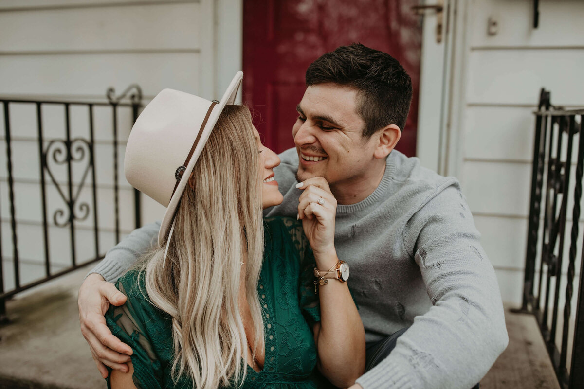 The couple sitting on the front steps of a house, sharing a loving gaze and smile, with the woman wearing a hat and the man in a sweater.