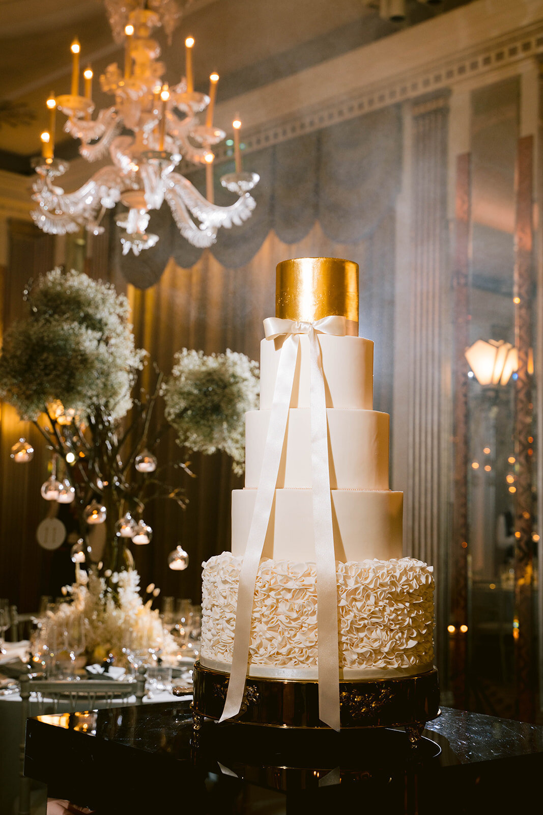 Wedding cake is in the Claridges ballroom with smoke in the background the cake has a gold tear a ruffle tear and a ribbon