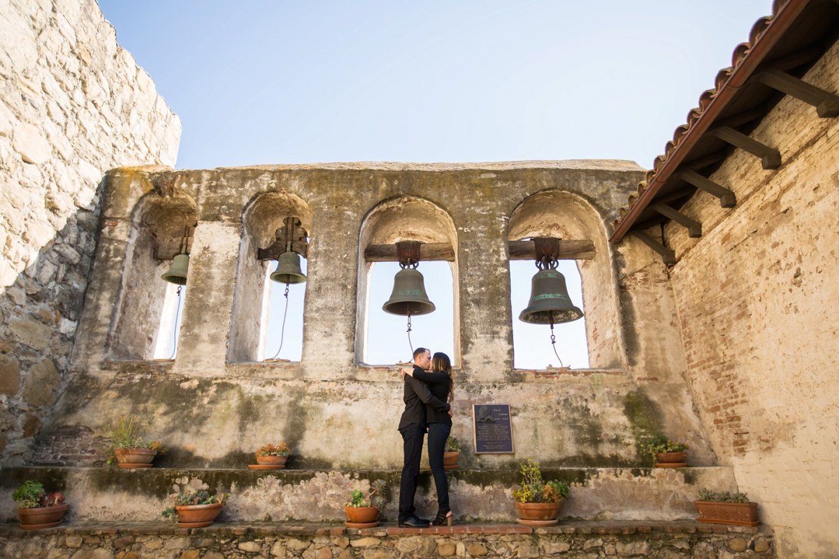 Engaged couple share a kiss in front of the bells at the Mission in San Juan Capistrano