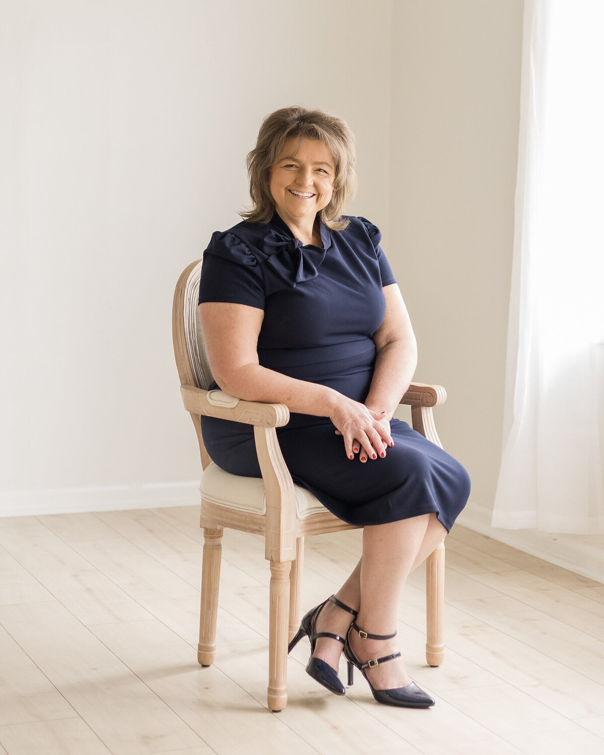 Brunette woman sitting in a chair with ankles crossed, wearing a navy blue dress, red nail polish, arms rested on one another, smiling at the camera