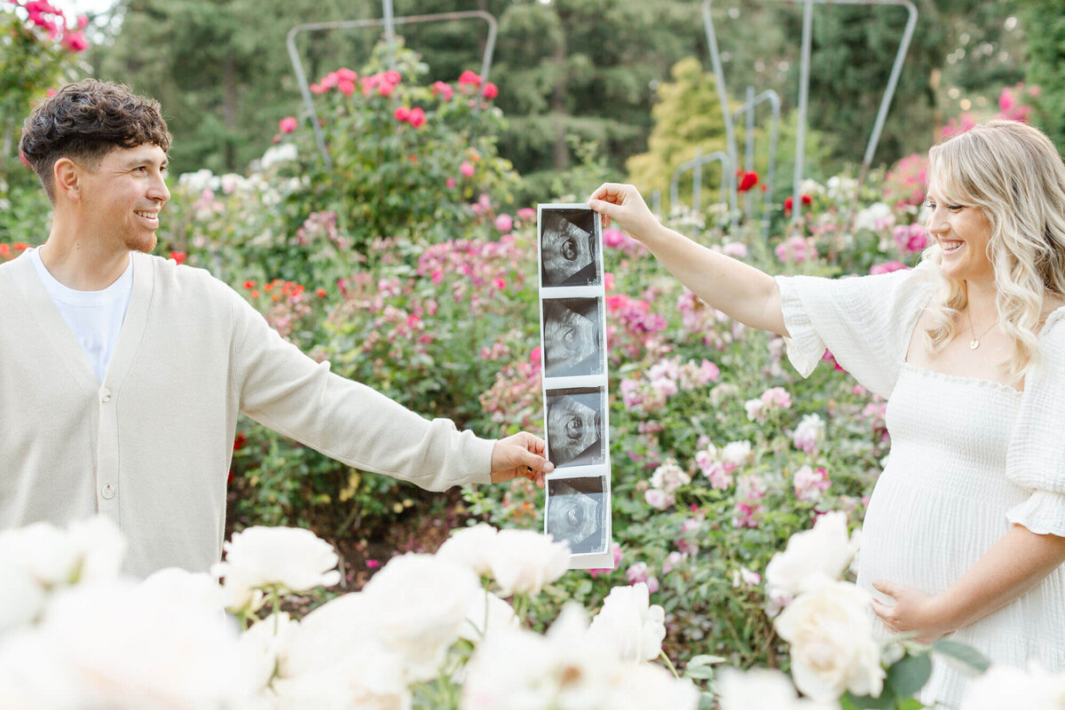 Pregnant Woman holding out her ultrasound photos and holding her belly, her husband is standing on the other side holding their ultrasound photos and they are both smiling at each other while standing in the middle of the Portland International Rose Test Gardens.