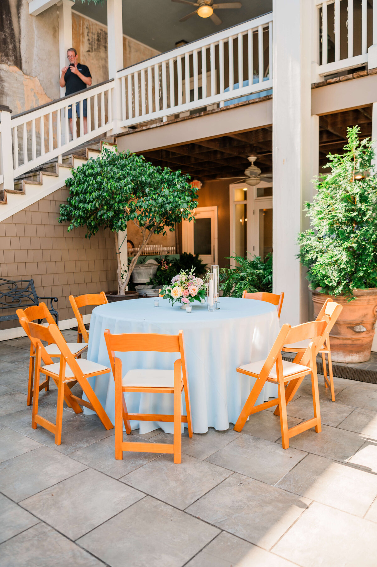 An outdoor courtyard set for an event, with round tables covered in white cloths, wooden chairs, floral centerpieces, potted plants, and string lights overhead.