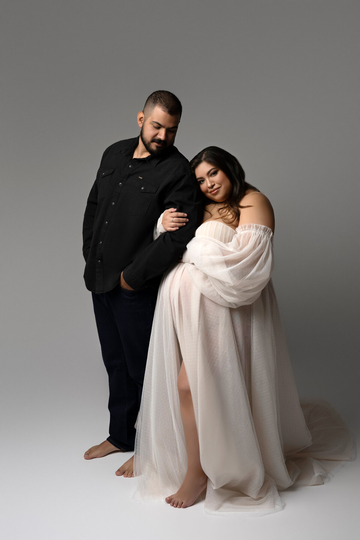 Romantic studio maternity portrait of a couple, with the mom-to-be in a flowy white off-shoulder gown and her partner in black.