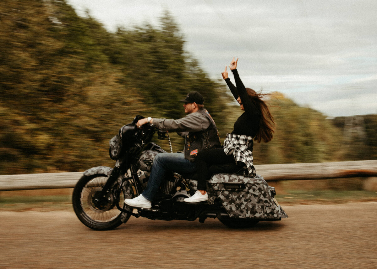 A guy is driving a motorcycle down the road while a girl sits behind him and throws her arms in the air.