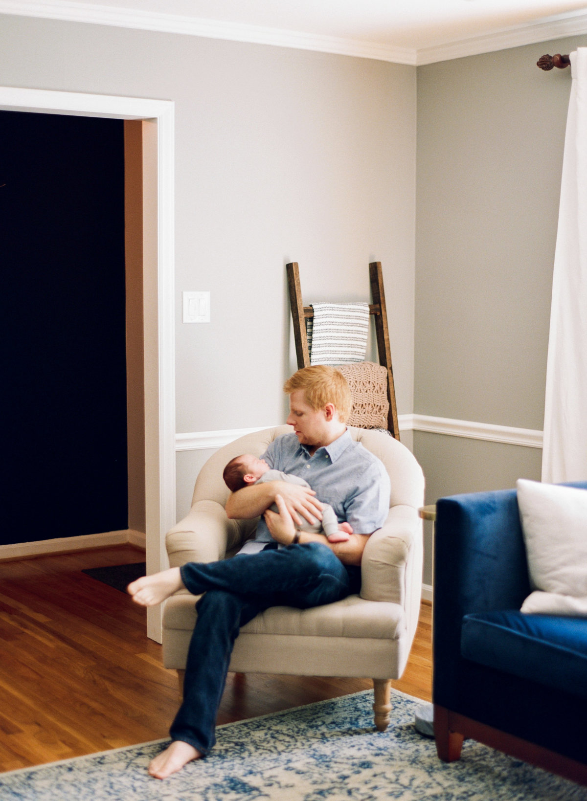 Dad sits in the living room looking adoringly at his newly adopted son during his newborn photography session in Raleigh. Photographed by Raleigh newborn photographers A.J. Dunlap Photography.
