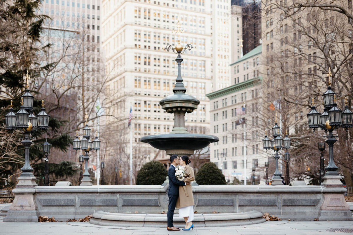 The bride and the groom are kissing in front of City Hall Park in NYC.