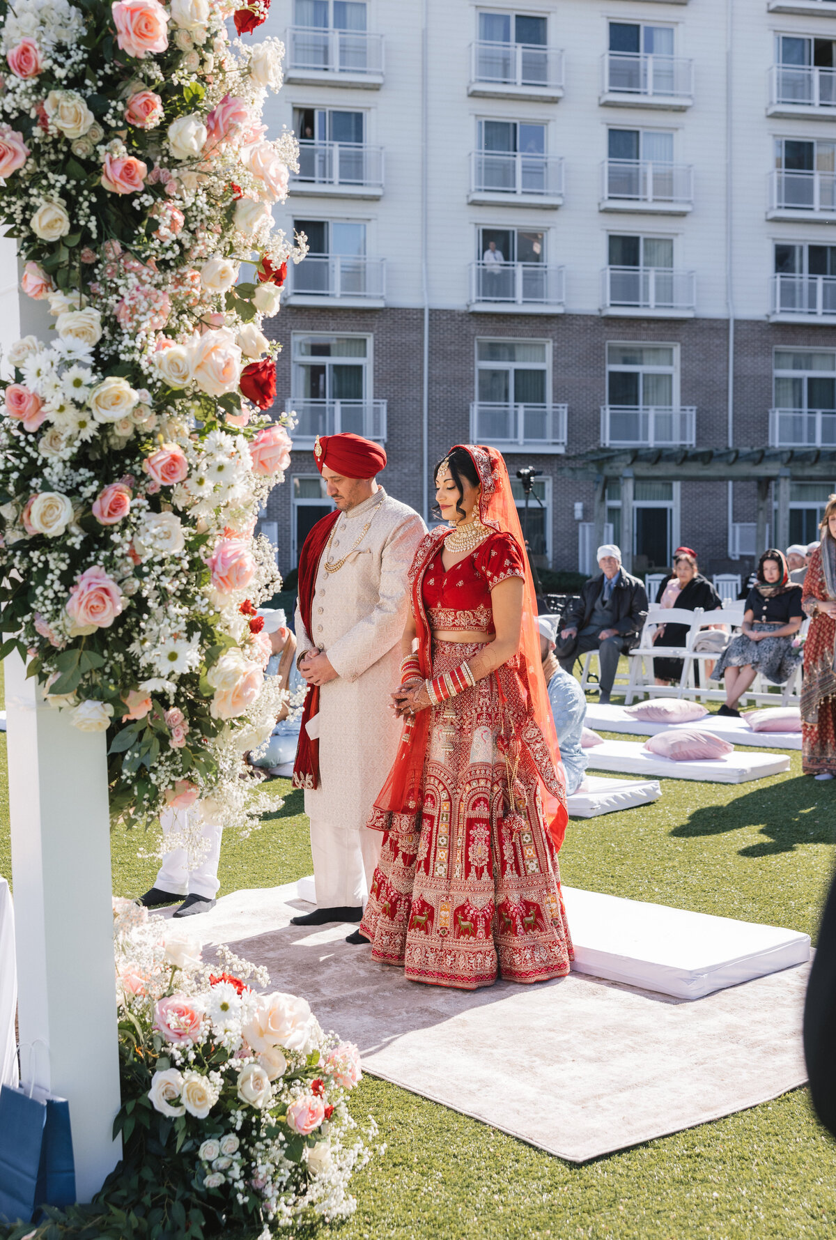 Bride and groom in red and cream attire posed beneath a floral outdoor mandap overlooking the water.