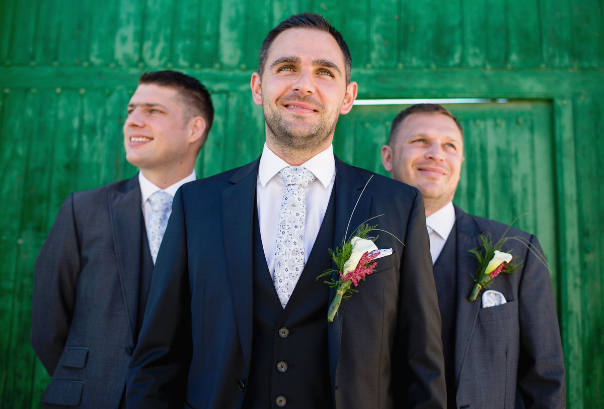 The Groom poses with his best man and usher behind him in front of a green wooden barn for a pre wedding photo taken by Motiejus wedding photographer while second shooting for Adorlee at this Spanish destination wedding.