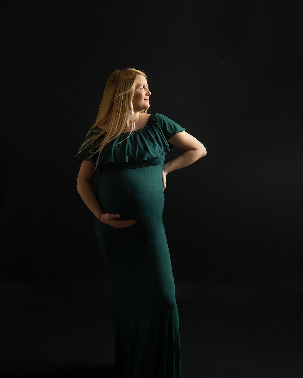 Pregnant woman in a green dress holding her baby bump with one arm and the other on her hip. The background is black.