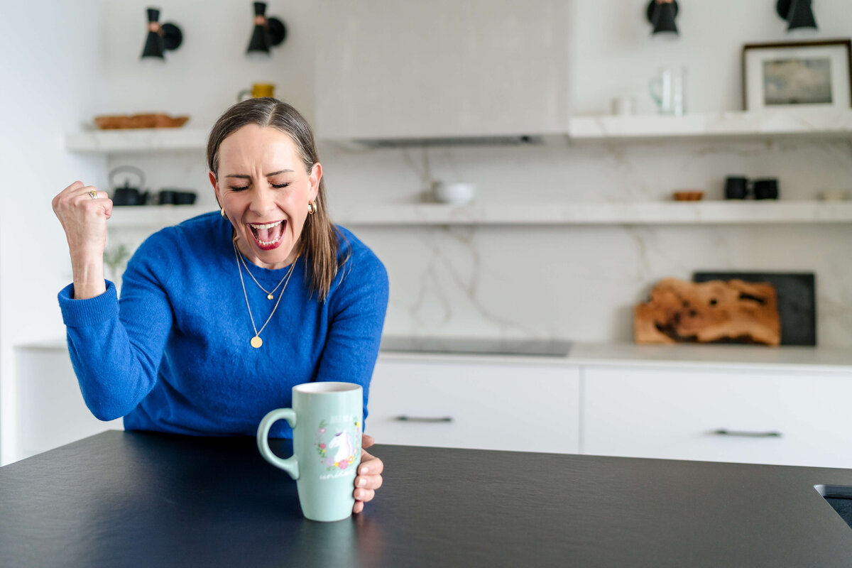 woman in blue sweater leaning on kitchen counter and holding green mug celebrating