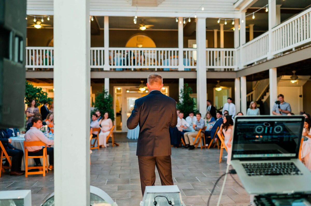 A man in a suit speaks to a seated audience at an outdoor venue, with a laptop open in the foreground and guests watching from tables and balcony areas.