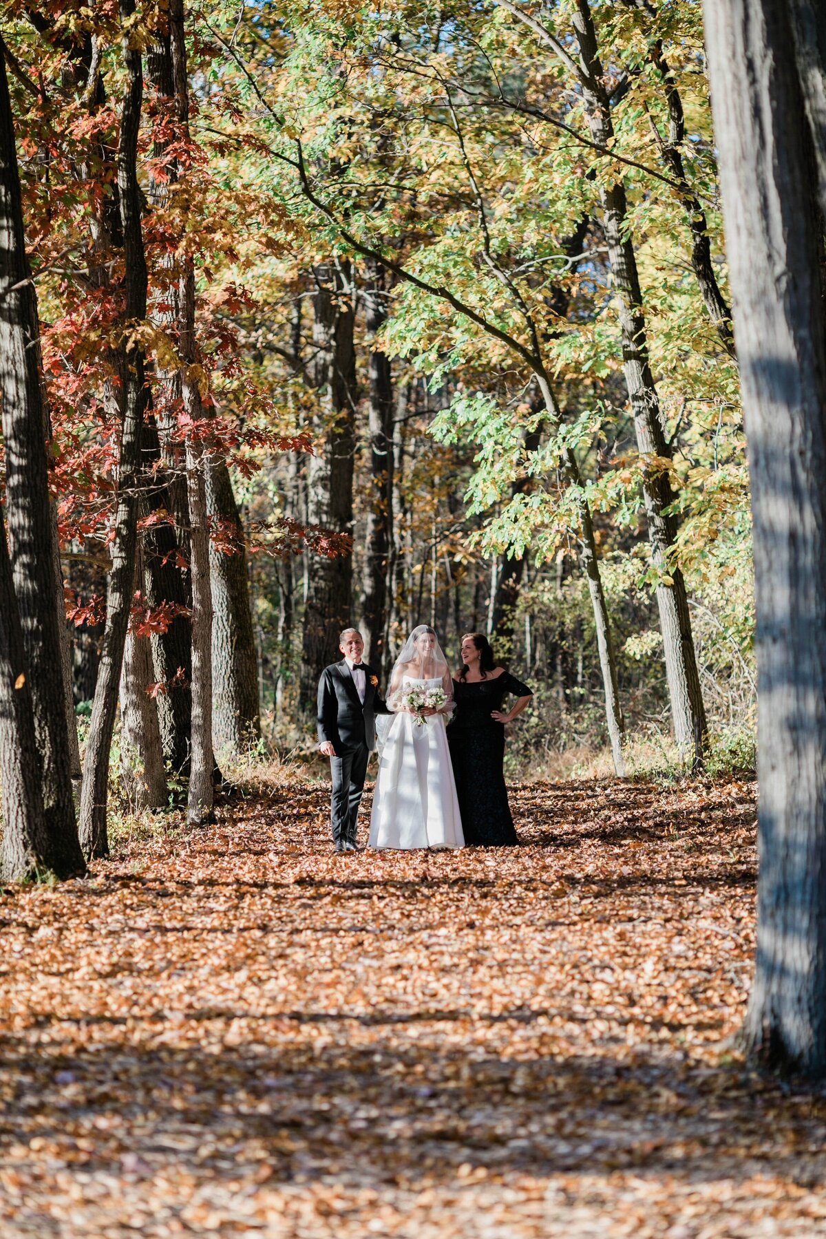 A bride, wearing a long veil and holding a bouquet, walks down a leaf-covered forest aisle