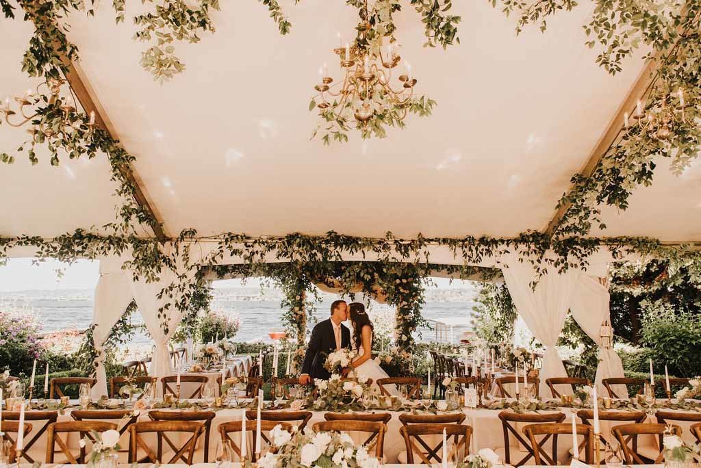 bride and groom in their wedding tent lined in ceiling greenery and long tables with vineyard chairs