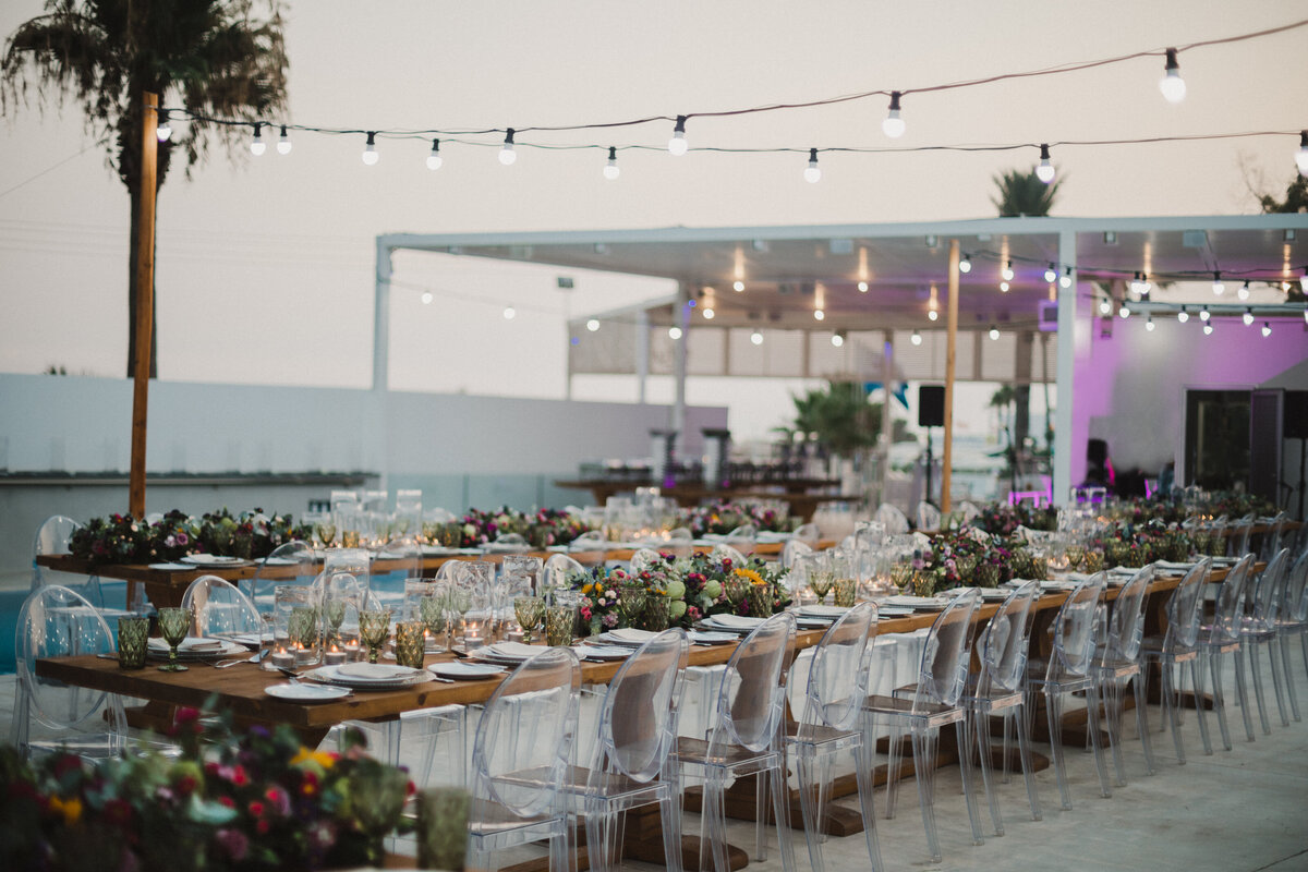 Long wodden tables are decorated with fresh flowers under festoon lighting