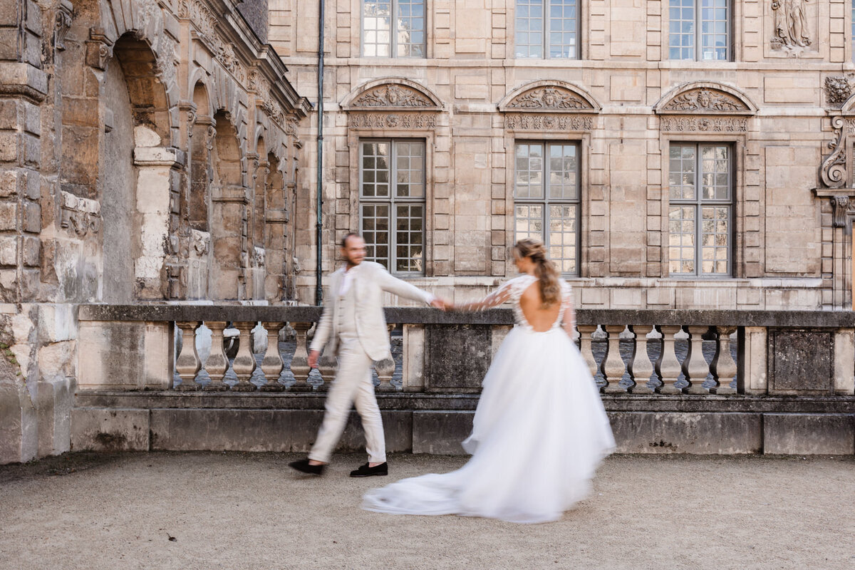Elopement parijs bruidspaar voor oud gebouw lopen elkaar voorbij raken elkaars handen aan en kijken om bruidegom in beige pak en bruid in addy van den krommenacker jurk