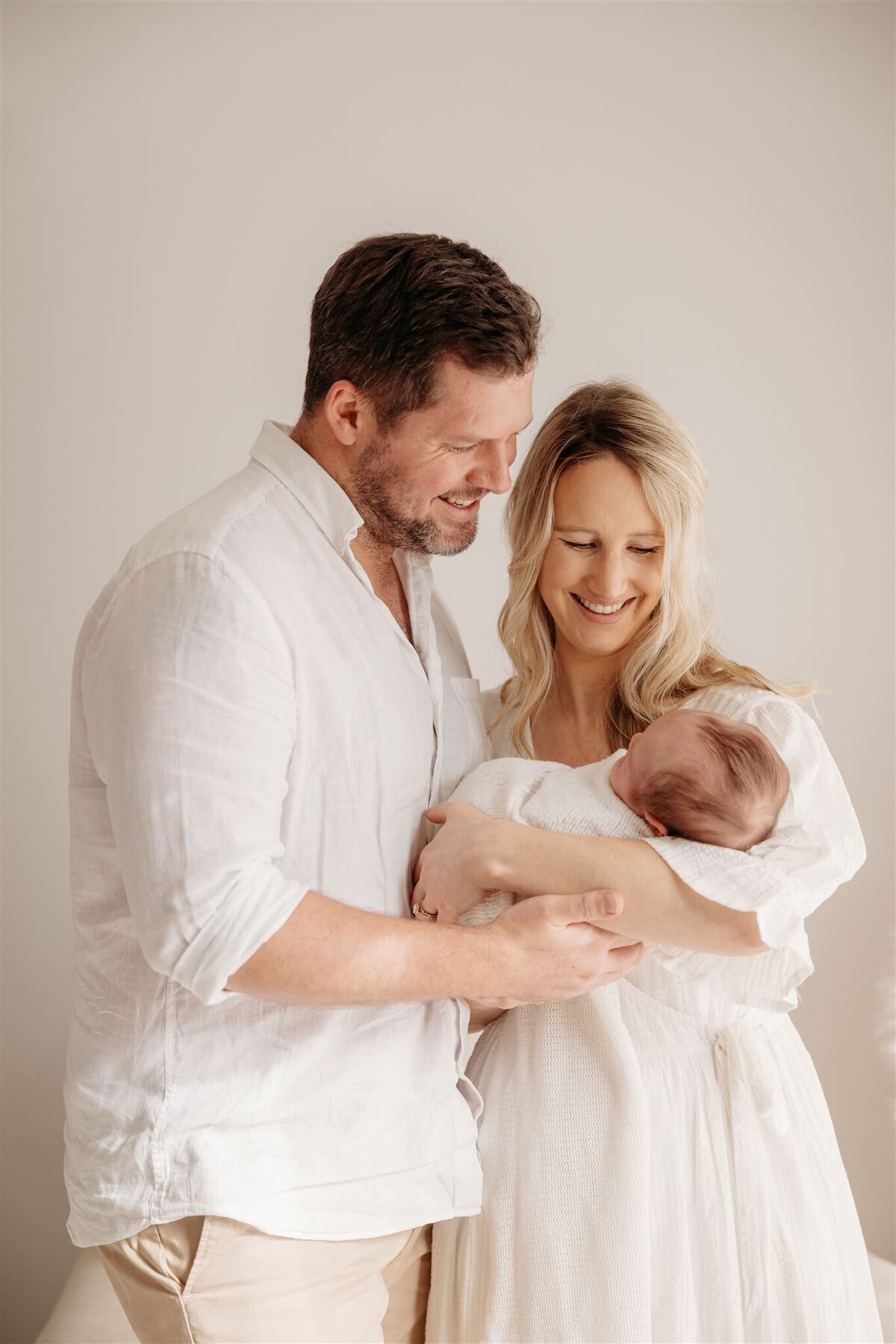 A mother and father holding their newborn baby all wearing white during a newborn photoshoot in auckland photography studio with michelle kelly photography