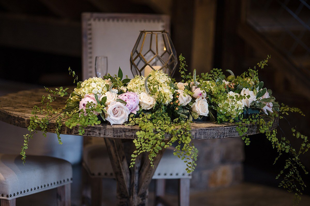 A round barn wood  farm table with studded white leather chairs is decorated with a tall geometric cut crystal lantern with draping greenery that cascades off the table accented with white roses, blush roses and ivory roses at L & L Farm.