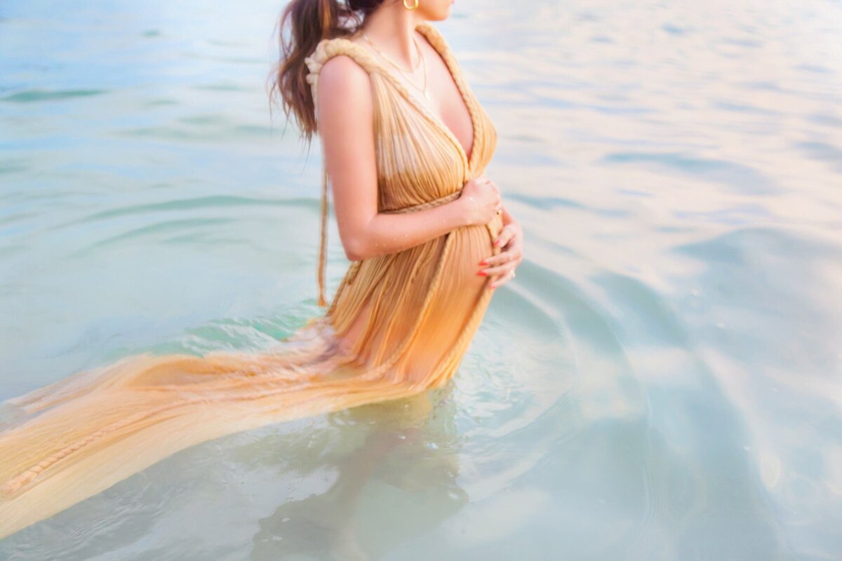 Woman in golden maternity gown holds her bump in the ocean while being photographed by Love + Water Photography during her babymoon in Wailea.