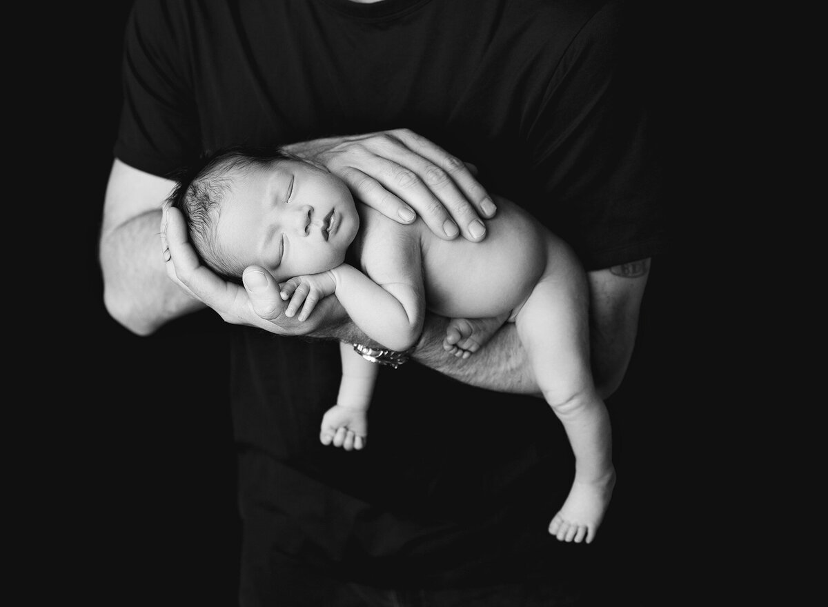 A father embraces his newborn, emphasizing the baby's tiny form set against a dark backdrop, capturing a tender moment.