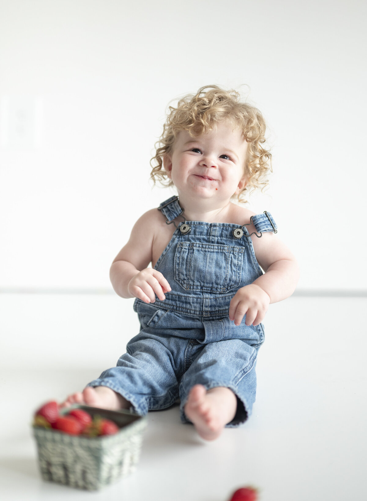 Laughing toddler in denim overalls  eating strawberries against a white background.