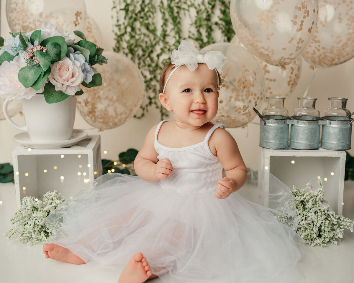 One year old girl in white tulle dress with white headband sitting on floor posing in front of balloon and floral decorations
