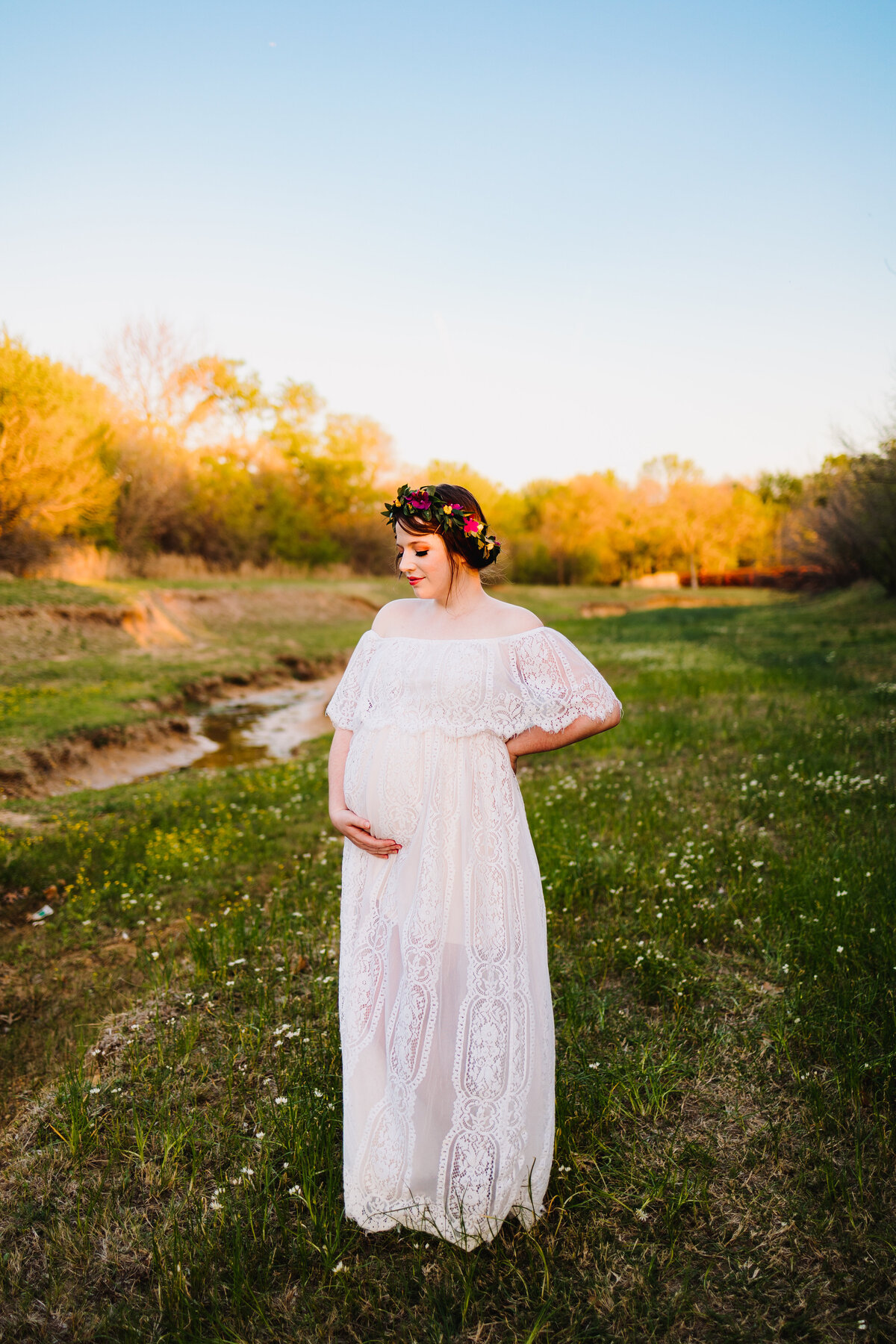 Photo of a pregnant woman in a forest with a small river behind, the woman has a long white dress and a headband of flowers with different colors