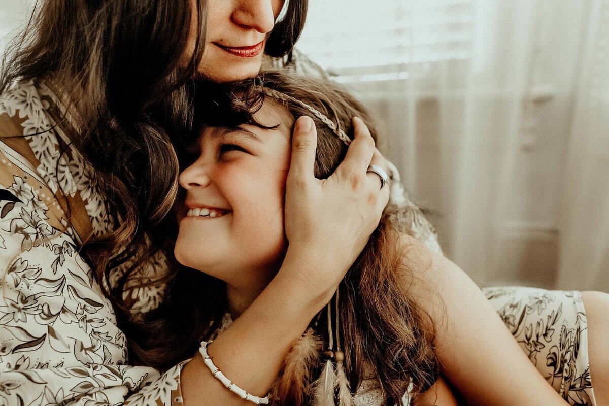 A woman embraces a smiling child in a warmly lit room. Both wear floral-patterned clothing.