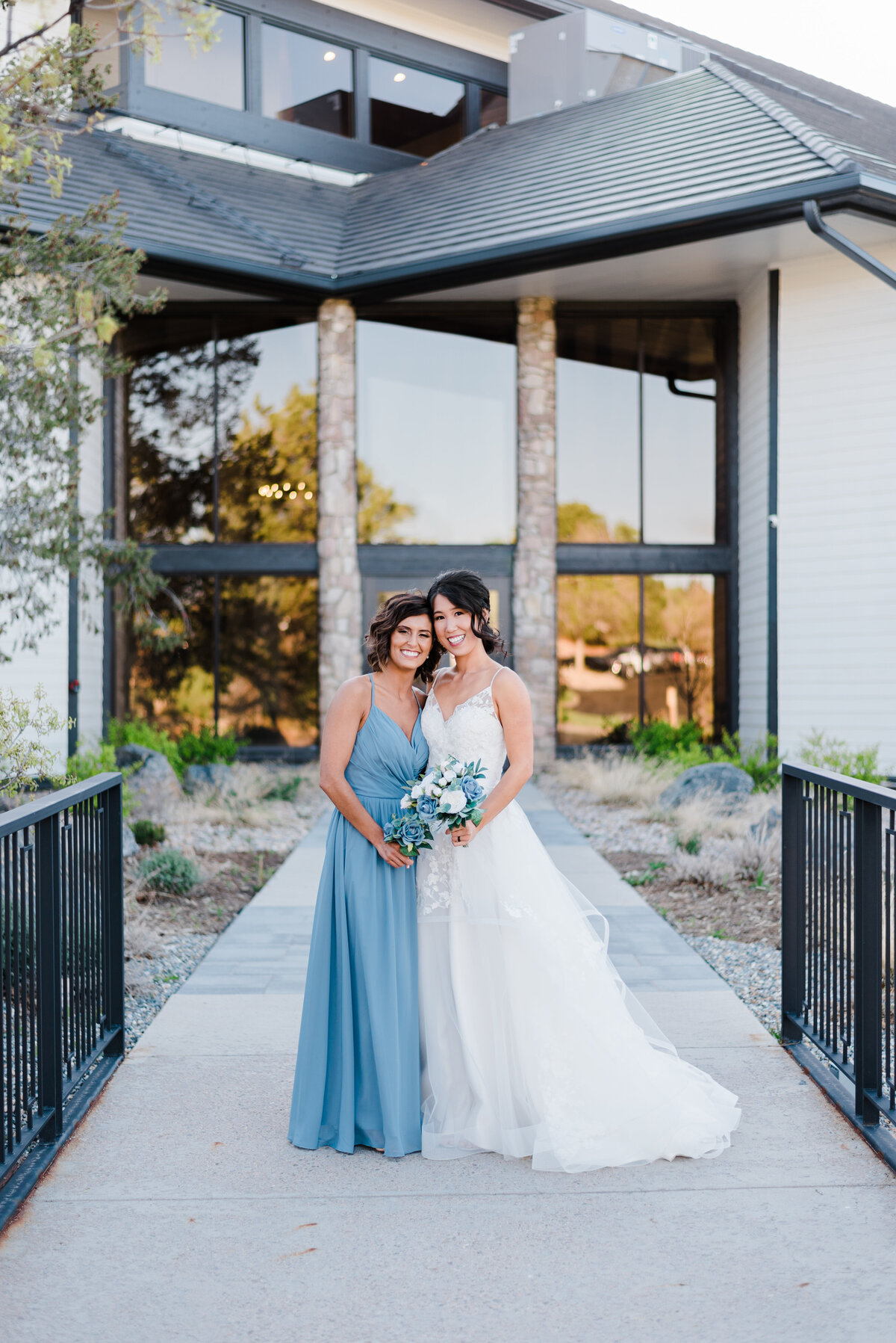 Bride and her maid of honor stand and hug outside The Oaks at Plum Creek