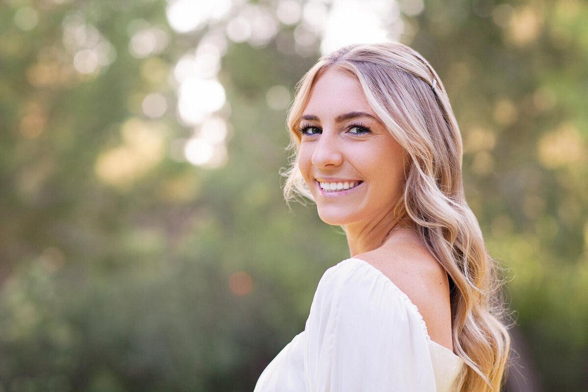 A woman with long, wavy blonde hair smiles at the camera. She is wearing a white off-the-shoulder top. The background is a blurred, green outdoor setting, suggesting a natural environment. The photo is well-lit, highlighting her features.