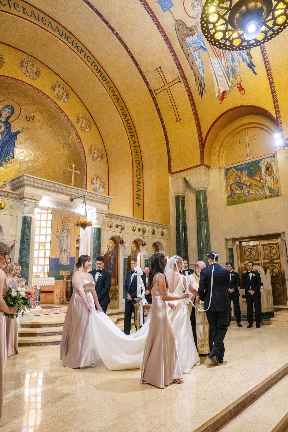 A couple stands at the altar during a wedding ceremony in an ornate, gold-adorned church. Bridesmaids in blush dresses adjust the brides train. Groomsmen in black suits stand nearby. Religious icons and arches are in the background.