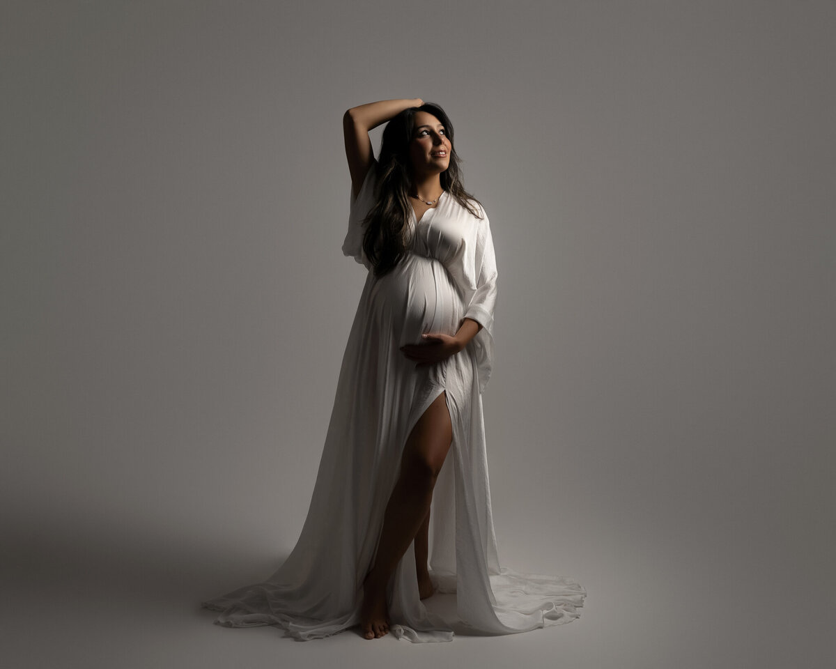 Elegant studio maternity portrait of a woman in a white robe with dramatic lighting and soft smile.