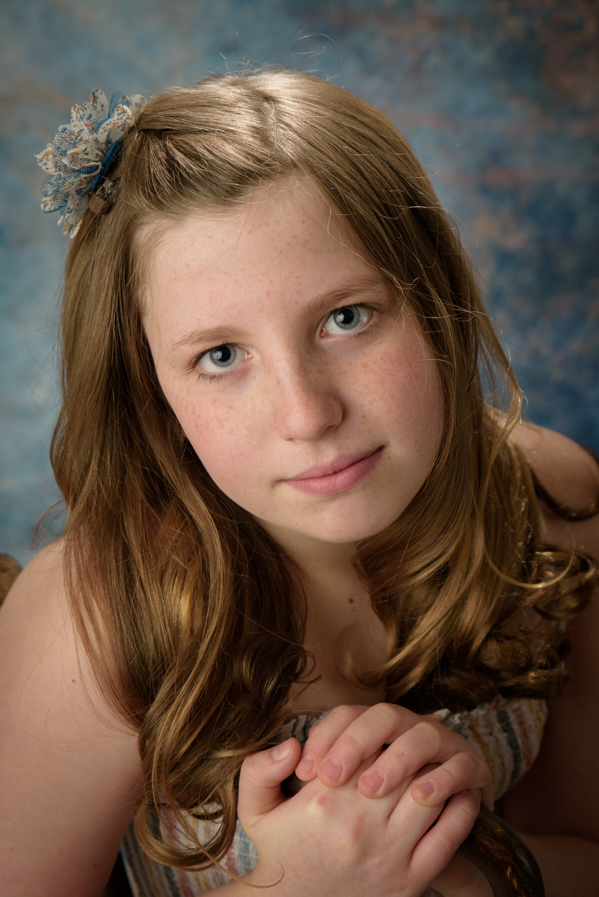 Beautiful young girl wearing a cream colored striped dress sitting on wooden chair in front of a blue background at my home studio near Green Bay, Wisconsin