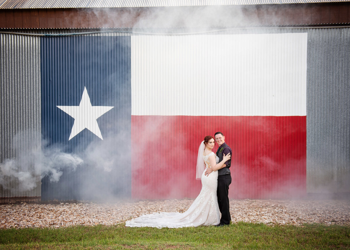 Bridal couple standing in front of a barn with the Texas flag painted on the side, at CW Hill Country Ranch.