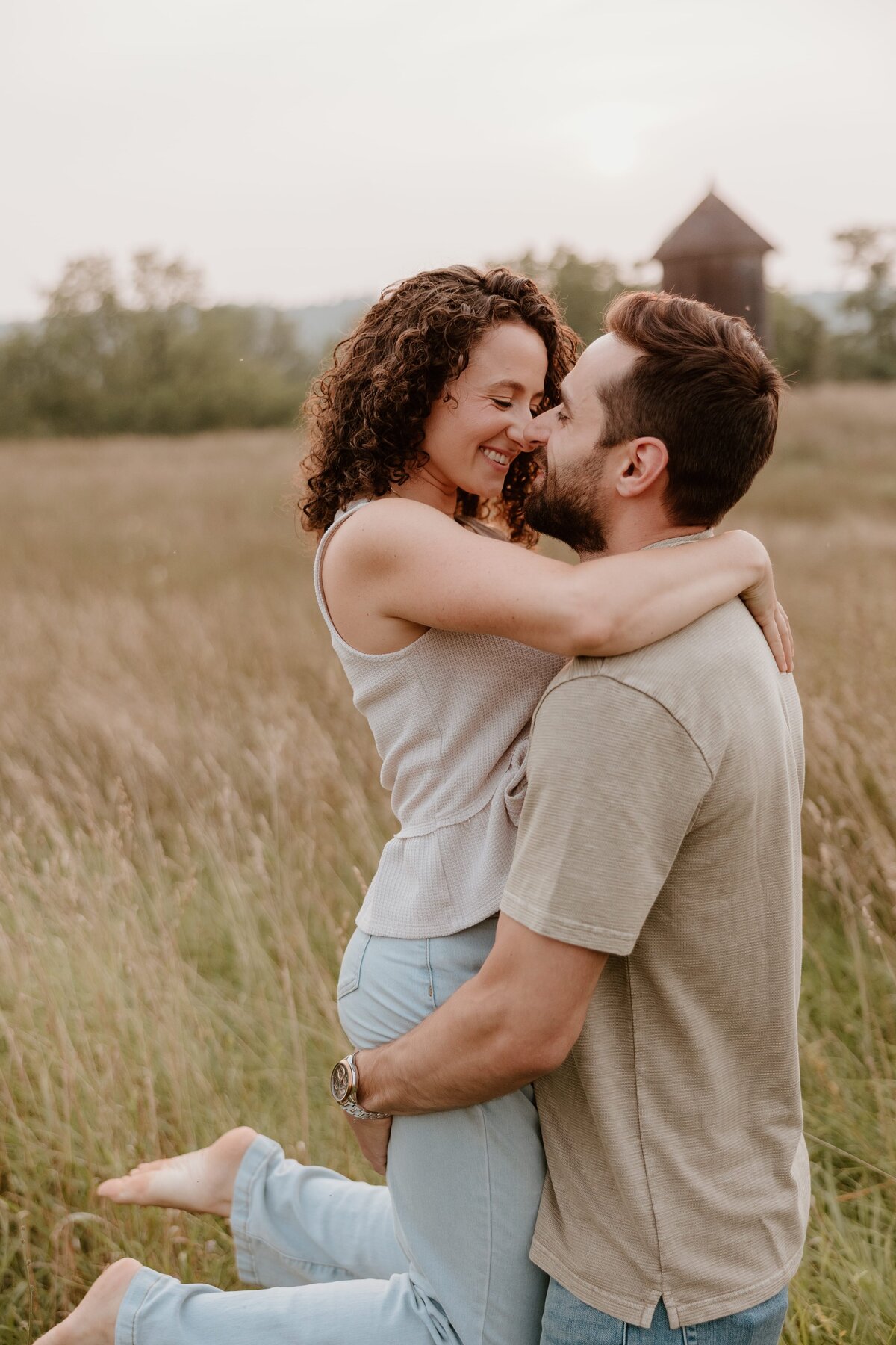 A joyful couple shares an intimate moment during their engagement session in a golden field at sunset, with the woman lifted in her partner’s arms and their noses touching.